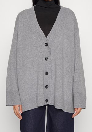 Persoon draagt een oversized lichtgrijze gebreide cardigan met knopen en geribde boorden over een zwarte coltrui en donkere broek.