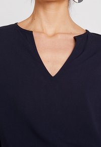 Nainen, jolla on päällään musta V-pääntiepaita, jossa on pieni lovi pääntien oikeassa sivussa, sekä suuret hopeiset korvarenkaat.