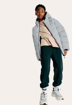 Giovane in giacca a vento grigia chiara, maglietta beige, pantaloni neri, calzini bianchi e sneaker bianche chunky, in piedi davanti a uno sfondo neutro.
