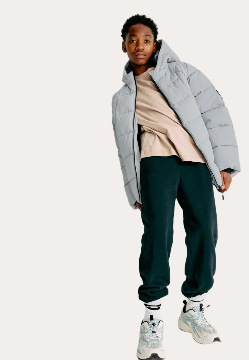 Ung person iført en lysegrå pufferjakke, beige skjorte, sorte sweatpants, hvide sokker og kraftige hvide sneakers, stående mod en neutral baggrund.
