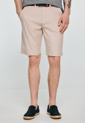 Mężczyzna w beżowych szortach do kolan, paskowanej koszuli, czarnym pasku i czarnych wsuwanych butach, stoi z rękami w kieszeniach.