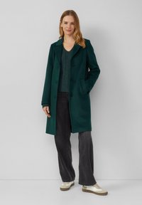 Dunkelgrüner Wollmischmantel mit Reverskragen, Knopfverschluss und zwei Taschen, kombiniert mit einem strukturierten grünen Pullover und dunkelgrauen Hosen.
