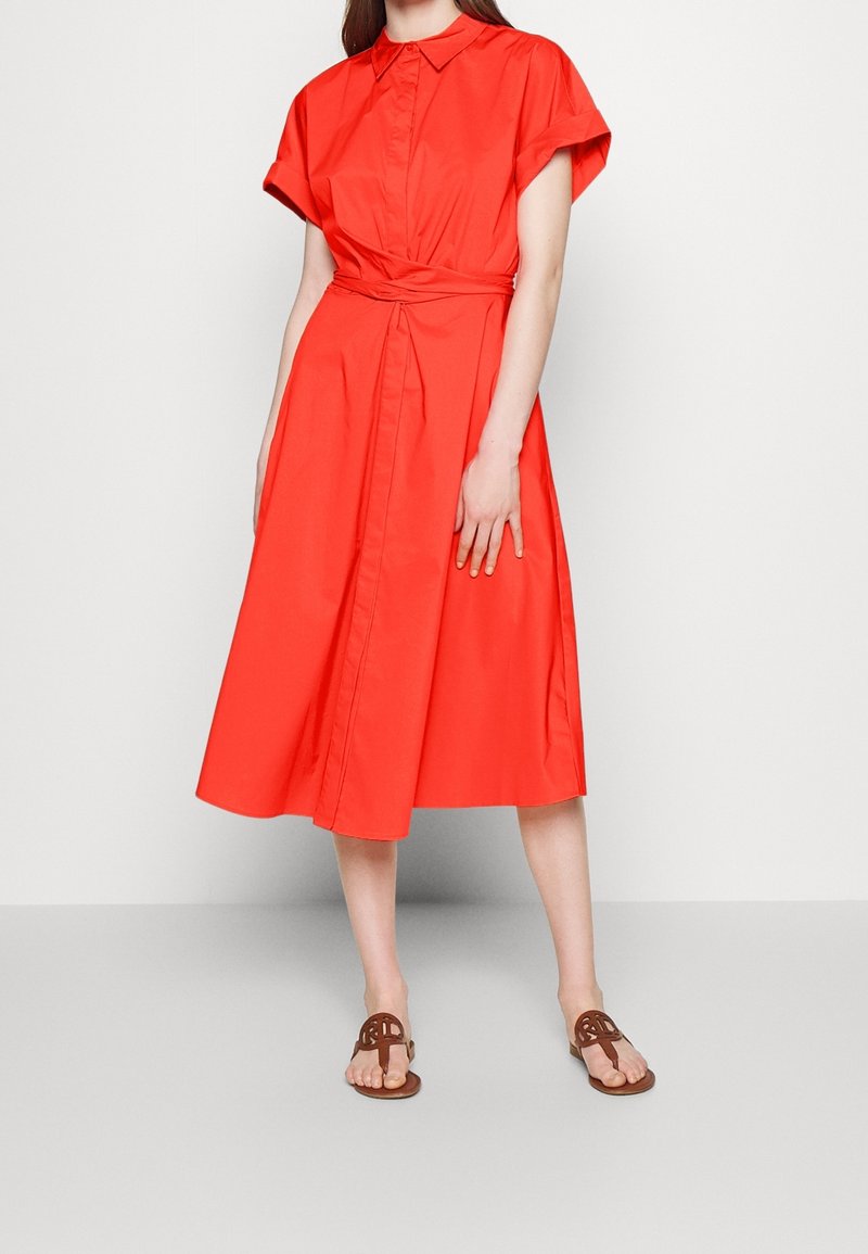 Kvinna iklädd en ljus röd, kortärmad midiklänning med midjeband och bruna öppna sandaletter, stående mot en enfärgad vit vägg.