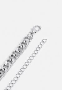 Bracelet en métal argenté avec une chaîne à maillons entrelacés, un fermoir mousqueton et une chaîne d'extension avec des maillons circulaires. Texture lisse et brillante.