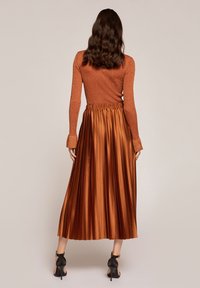 Bronzový žebrovaný top s dlouhým rukávem v kombinaci s plisovanou saténovou maxi sukní ve podobném odstínu. Černé kotníkové boty na podpatku dokončují vzhled.