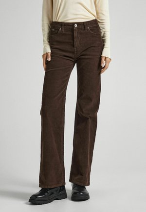 Osoba ubrana w ciemnobrązowe spodnie sztruksowe o prostych nogawkach, kremową bluzkę z długim rękawem z postrzępionymi mankietami oraz czarne masywne buty stojąca na szarej podłodze.