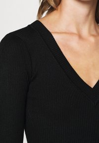 Svart ribbad stickad topp med djup V-ringning, med en texturerad tyg och figurnära design. Närbild av ringningen och axeln.