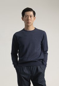 Jersey de punto azul marino con mangas largas y cuello redondo, que presenta un sutil detalle de logotipo. Combinado con pantalones oscuros con pliegues.