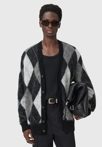 Cardigan argyle noir et gris, texture tricotée, coupe oversize, six boutons, porté avec un haut noir et tenant un sac en cuir noir.
