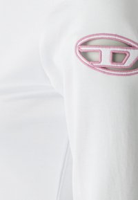 Hvit langermet skjorte med teksturert stoff. Merkbar rosa brodert logo på venstre erme med utskåret detaljering.