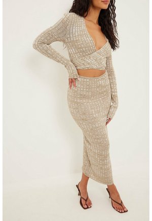 Nainen beigeä ribbineulosta kaksiosaisessa asussa, jossa on pitkähihaisen crop topin ja korkeavyötäröisen midihameen yhdistelmä, yhdistettynä ruskeisiin korkokengällisiin sandaaleihin.