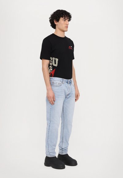 T-shirt noir avec design graphique, jeans bleu clair et chaussures noires. Caractéristiques : col rond et manches courtes, avec des éléments contrastants sur le t-shirt.