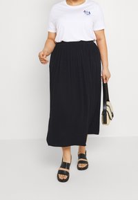 Persona che indossa una maglietta bianca a maniche corte, una gonna midi nera plisè, sandali neri e tiene in mano una piccola borsa beige e nera.