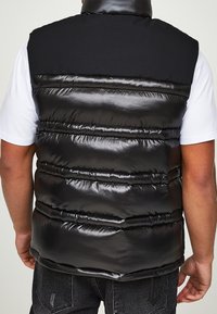 Mann trägt eine glänzende schwarze gesteppte Steppweste über einem weißen T-Shirt, von hinten gezeigt, mit dunklen Jeans.