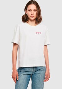T-shirt bianco in cotone con una vestibilità rilassata, caratterizzato da tre contorni di cuori rosa sul lato sinistro del petto. Maniche corte e scollo rotondo.