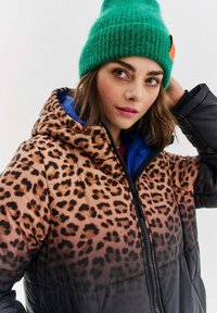 Žena v zelenej pletenej čiapke a leopardím ombré bomberi s modrým vnútorným podšívaním, ktorá jednou rukou upravuje čiapku.