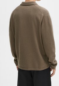Homme portant un pull en maille marron à manches longues et un pantalon noir, debout de dos devant un fond blanc uni.