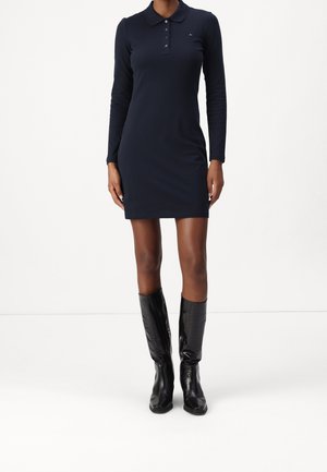 Femme portant une robe polo longue à manches longues bleu marine, associée à des bottes en cuir noir texturé montant jusqu'aux genoux, se tenant devant un fond blanc uni.