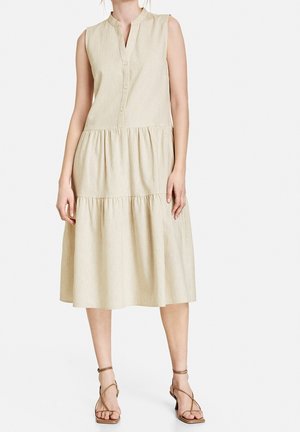 Robe midi beige sans manches avec col en V boutonné et jupe à volants, portée avec des sandales à talons bas et brides sur un mannequin sur fond blanc.