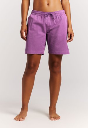 Persoon die blootsvoets staat, gekleed in paarse mid-dij korte broek met een elastische tailleband en trekkoord, tegen een effen lichte achtergrond.