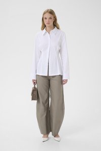 Woman wearing white buttoned shirt, wide-leg taupe pants, white pointed shoes, holding taupe handbag, standing against plain background.