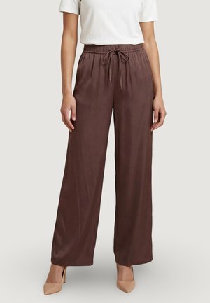 Žena stojící proti jednoduchému světlému pozadí, oblečená v širokých hnědých saténových kalhotách se stahovacími šňůrkami v pase a béžových vysokých podpatcích.