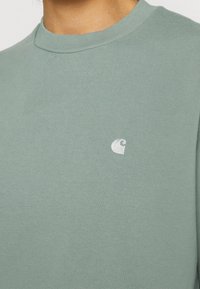 Gros plan d'une personne portant un t-shirt vert clair à col rond avec un petit logo blanc sur la poitrine.