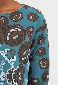 Mönstrad blus i flera färger med en turkos bakgrund och bruna och vita blommönster, rund halsringning och mjuk tygkänsla.