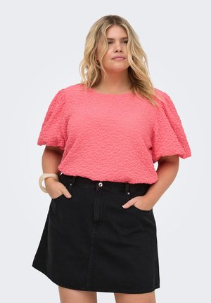Femme aux cheveux blonds ondulés portant un haut texturé corail rose à manches courtes et une jupe en jean noire, mains dans les poches, sur fond gris clair.