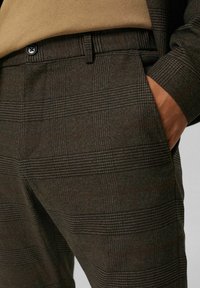 Gros plan d'une personne portant un pantalon à carreaux marron foncé avec une ceinture boutonnée et la main dans la poche latérale.