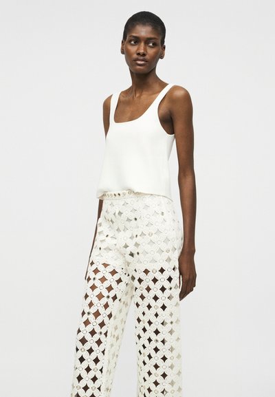 Femme portant un haut blanc sans manches et un pantalon large blanc avec un motif floral découpé, debout devant un fond clair uni.