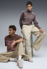 Două modele îmbrăcate în ținute sport casual: o jachetă mauve cu fermoar, un tricou grafic maro, pantaloni largi bej deschis și adidași roz de la Puma.