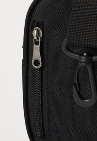 Zwarte stoffen zak met ritssluiting en zilveren ritshardware. Beschikt over een stevige strapbevestiging en een gestructureerd oppervlak voor extra grip.