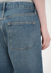 Gros plan sur l'arrière d'un jean en denim bleu avec une poche visible et une main reposant à côté de la hanche.