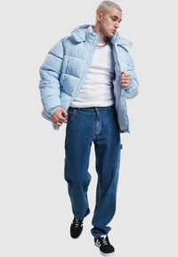 Lys blå dunsjakke med hette, med quiltede paneler, glidelås foran og ribbede mansjetter, kombinert med løstsittende blå jeans og svarte sneakers.