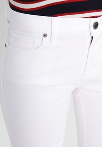 Jeans de mezclilla blanca con un diseño ajustado, bolsillos frontales y cierre de botón. La tela tiene una textura suave con un estiramiento mínimo.