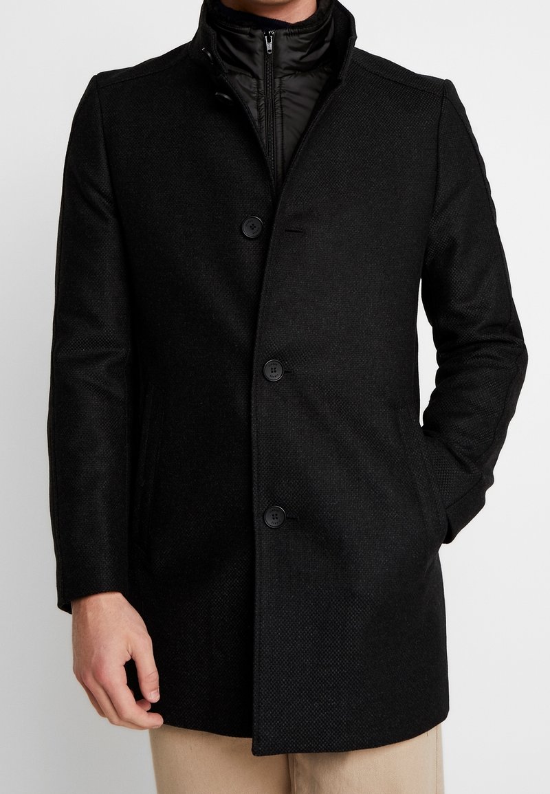 Manteau noir avec un motif texturé subtil, présentant un col haut, six boutons et des poches latérales. L'intérieur révèle une couche matelassée noire.