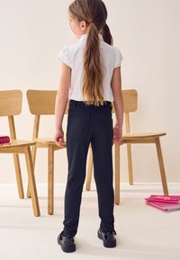 Musta slim-fit-housut, joissa on takataskut, yhdistettynä lyhythihaiseen valkoiseen paitaan. Lapsi seisoo kahden puutuolin edessä.