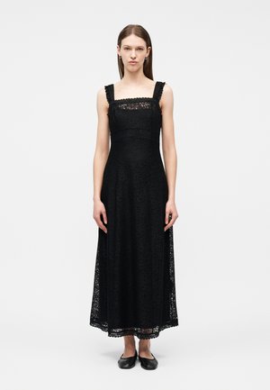 Vrouw staand, draagend een zwarte kanten mouwloze midi-jurk met een vierkante halslijn en zwarte platte schoenen, op een witte achtergrond.