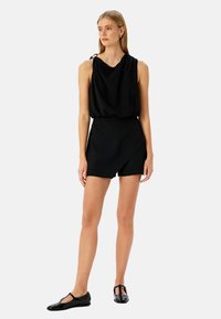 Top negro sin mangas con tela drapeada y escote bandeja, combinado con pantalones cortos negros ajustados. La modelo lleva zapatos planos negros.