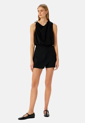 Top negro sin mangas con tela drapeada y escote bandeja, combinado con pantalones cortos negros ajustados. La modelo lleva zapatos planos negros.
