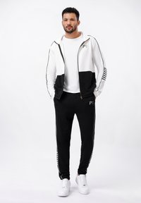 Heren trainingspak met een wit en zwart jack met ritssluiting, voorzien van een bedrukte logo, gecombineerd met zwarte joggers, afgewerkt met witte strepen.