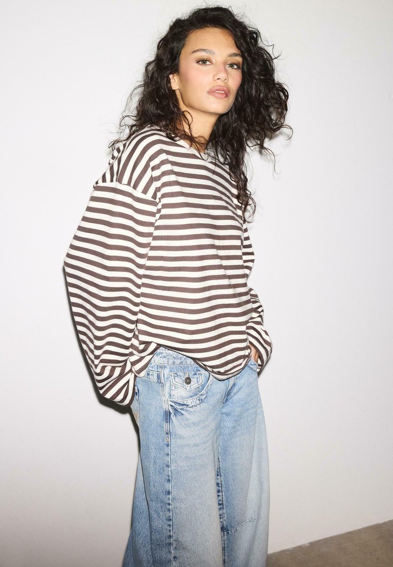 Junge Frau mit lockigem dunklem Haar, die ein braun-weiß gestreiftes übergroßes Hemd und hellblaue Jeans mit weitem Bein trägt, steht vor einer weißen Wand.