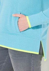 Felpa blu chiaro con tasca frontale, caratterizzata da un orlo spaccato e trim giallo neon lungo i polsini e il bordo inferiore.