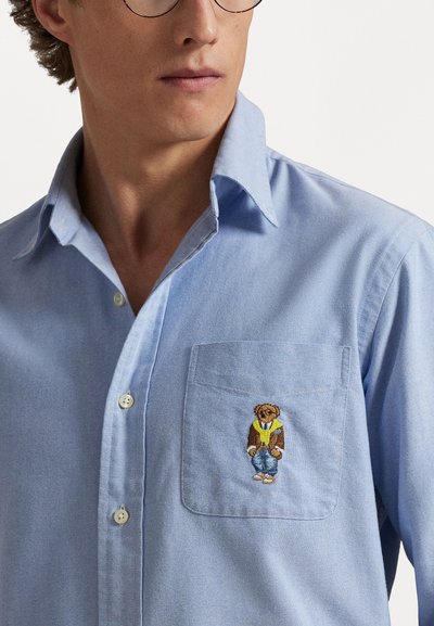 Gaiši zila krekla ar pogām dizains, kas piedāvā krūšu kabatu ar izšūtu brūnu lāci ikdienas apģērbā. Materiāls izskatās gluds un austa.