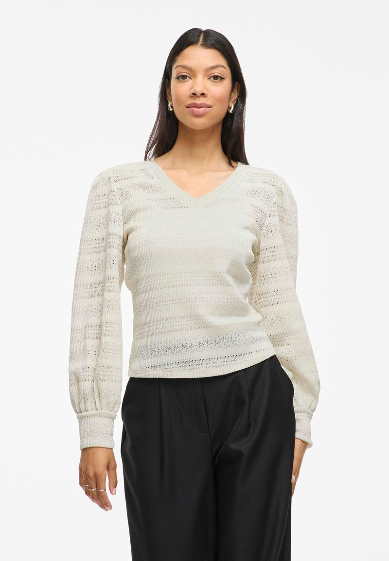 Light beige pullover top with a V-neck, textured stripes, and puffed long sleeves. Paired with loose black pants.