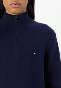 Granatowy sweter z wysokim kołnierzem i ćwierć zamkiem. Posiada małe logo w kolorach czerwonym, białym i niebieskim po lewej stronie klatki piersiowej. Miękka tekstura.