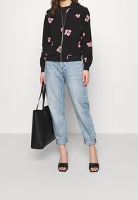 Svart zip-up bomberjacka med rosa blommönster, i kombination med ljusblå uppvikta jeans. Modellen håller en svart tote-väska och bär svarta sandaler.