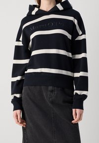 Sudadera con capucha de rayas blancas y negras con un logo en el frente, hecha de tela suave. Cuenta con un bolsillo tipo canguro y puños y ribetes acanalados.