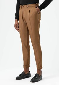 Bruine op maat gemaakte broek met een geplooide voorkant, vervaardigd uit een textuurstof. Draagt zwarte loafers en een zwarte trui.